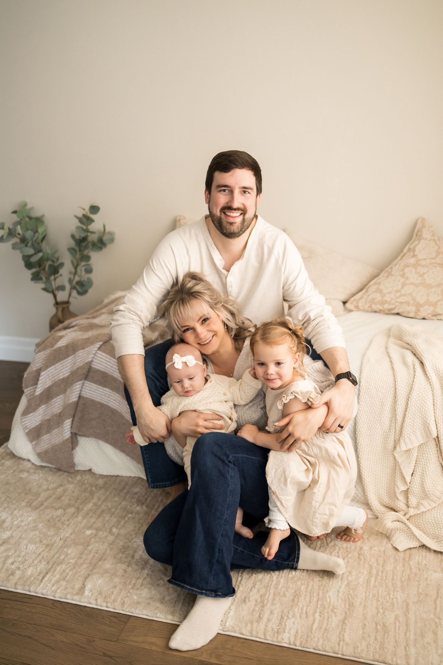 Family in cream-colored outfits lounging together on beige carpet and bedding for casual indoor photoshoot.