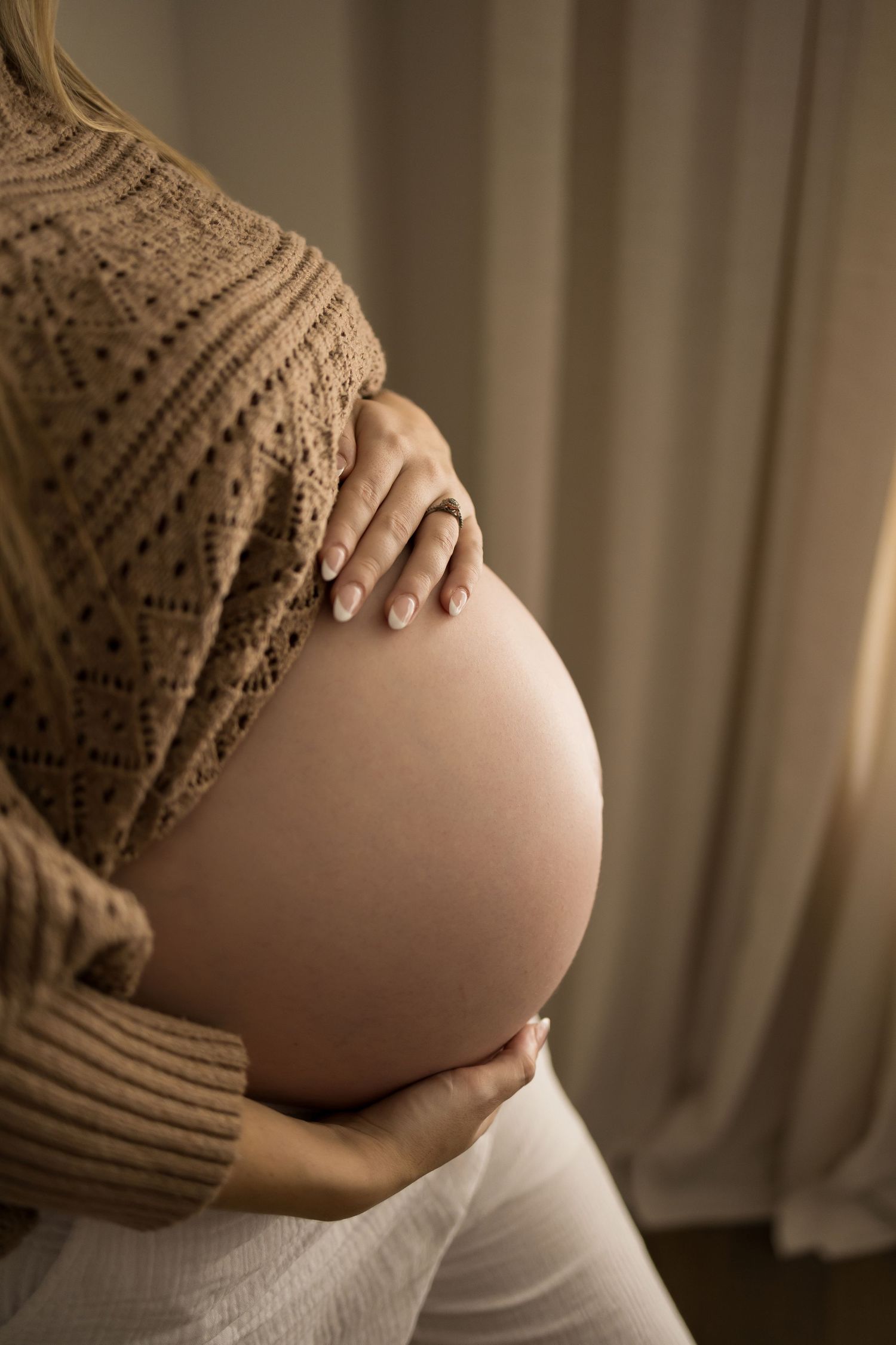 A pregnant person wearing a brown knit sweater gently cradles their belly in a softly lit room.