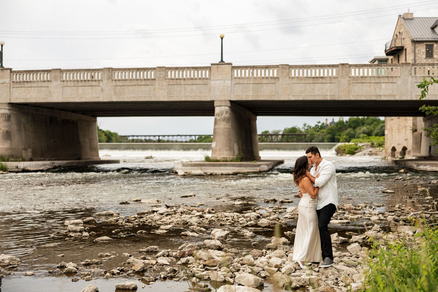 Bride and groom embrace on rocky riverbank beneath elegant stone bridge on their wedding day.