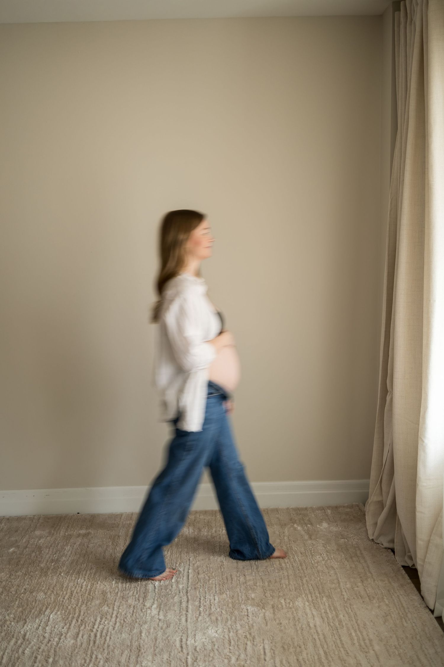 Pregnant woman in jeans and white top walking in a minimalist room, captured with motion blur effect.