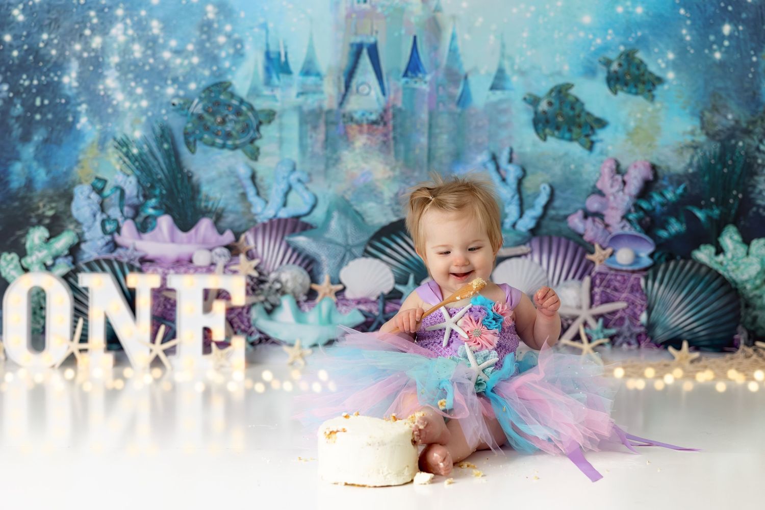Baby in tulle dress celebrates first birthday with cake against magical underwater backdrop.