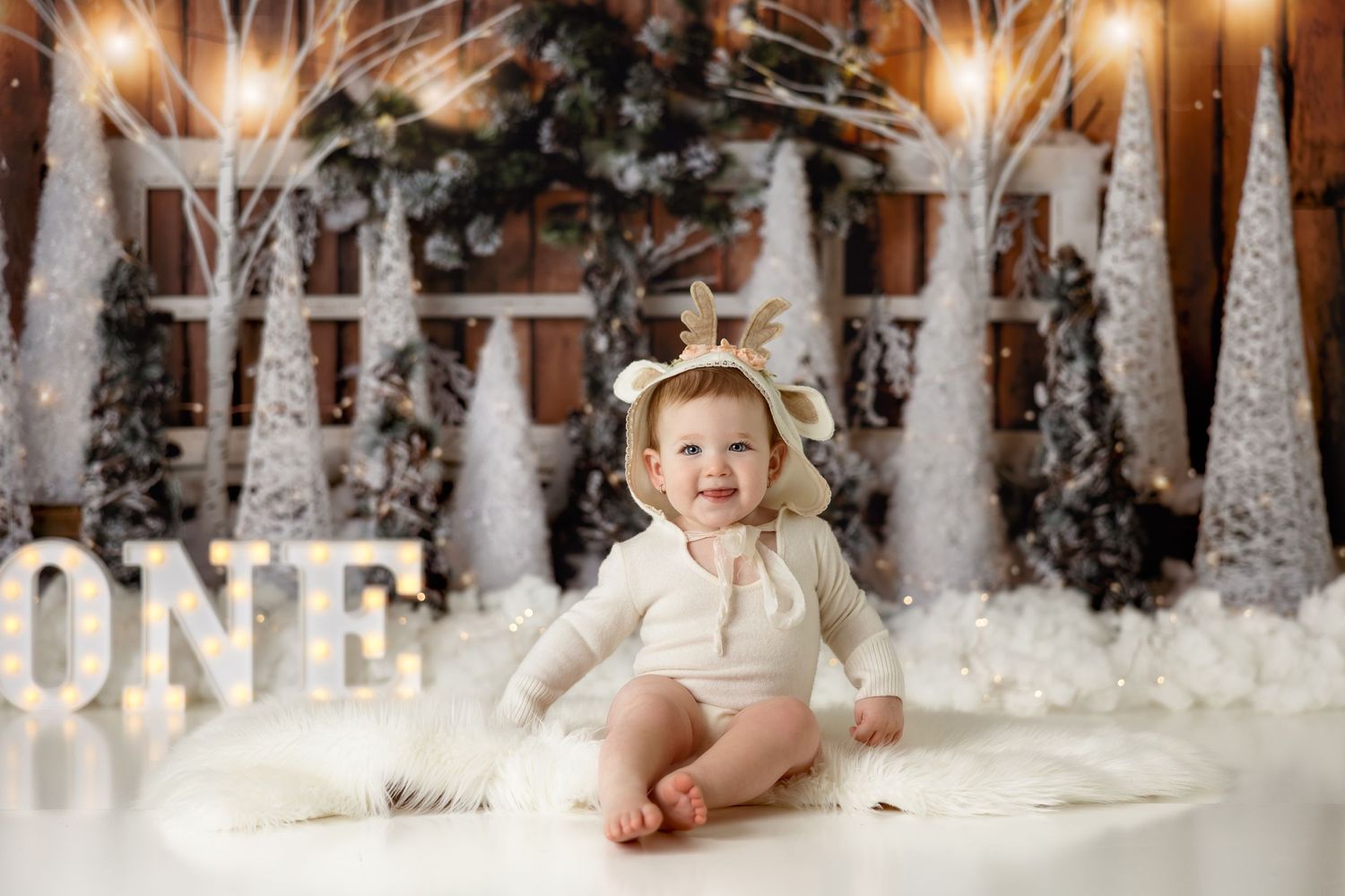 Baby in cream colored outfit sits on floor with winter forest scene and ONE sign backdrop.