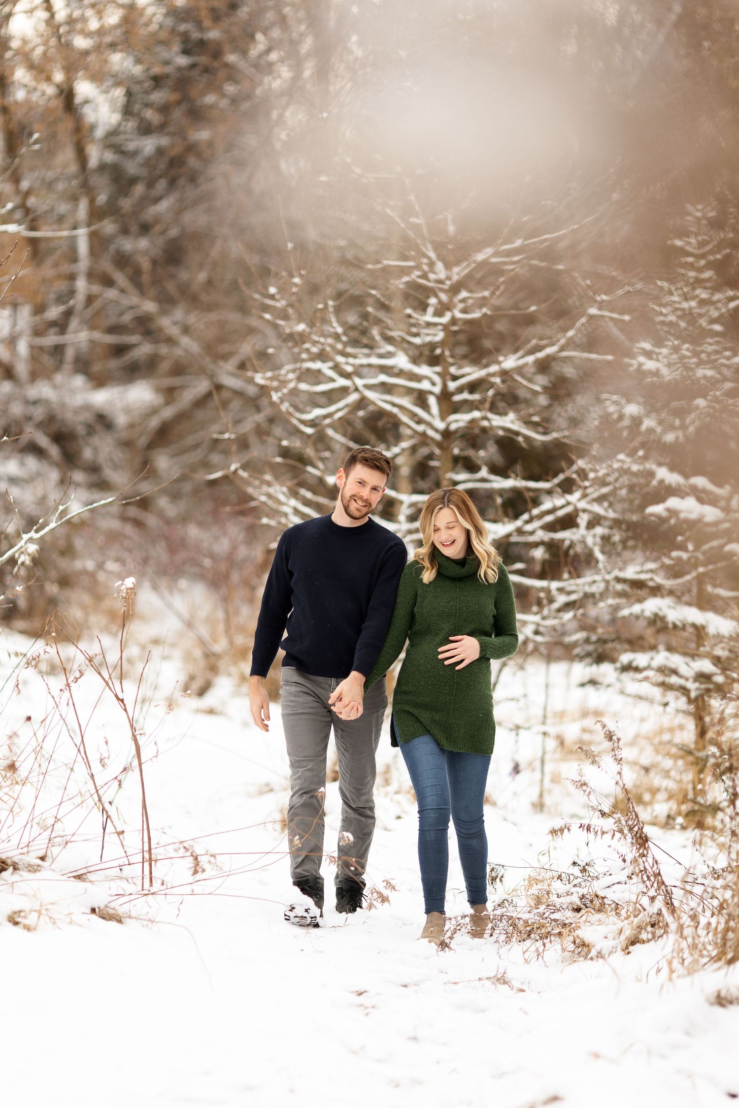 A winter maternity photo of a couple walking through snow-dusted trees.