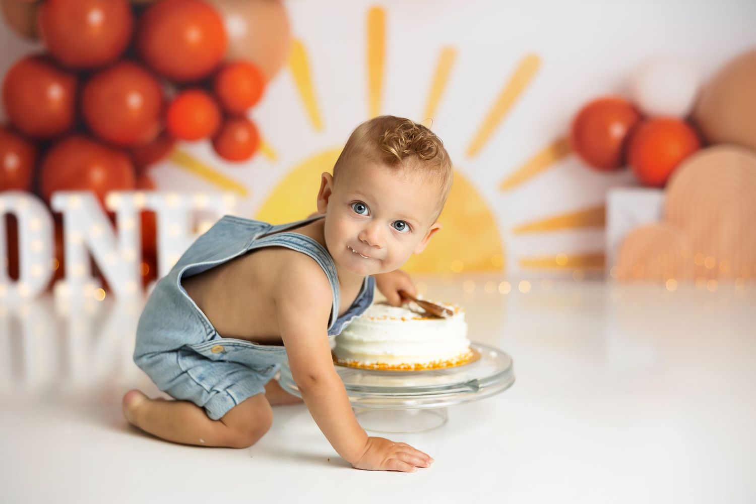 One-year-old in blue romper crawls toward birthday cake with orange balloon display.