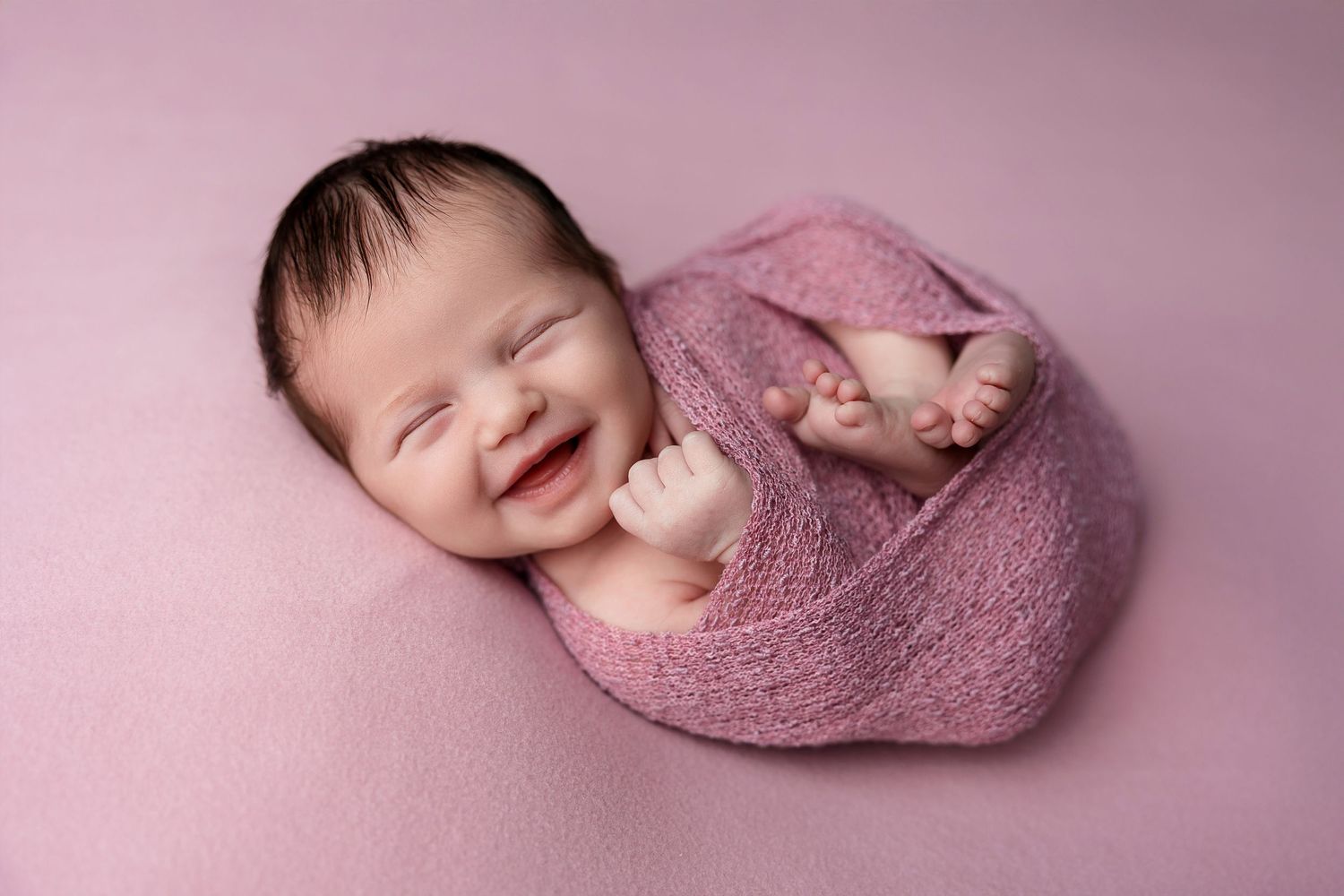 A newborn wrapped in pink fabric smiles while sleeping on a pink backdrop.