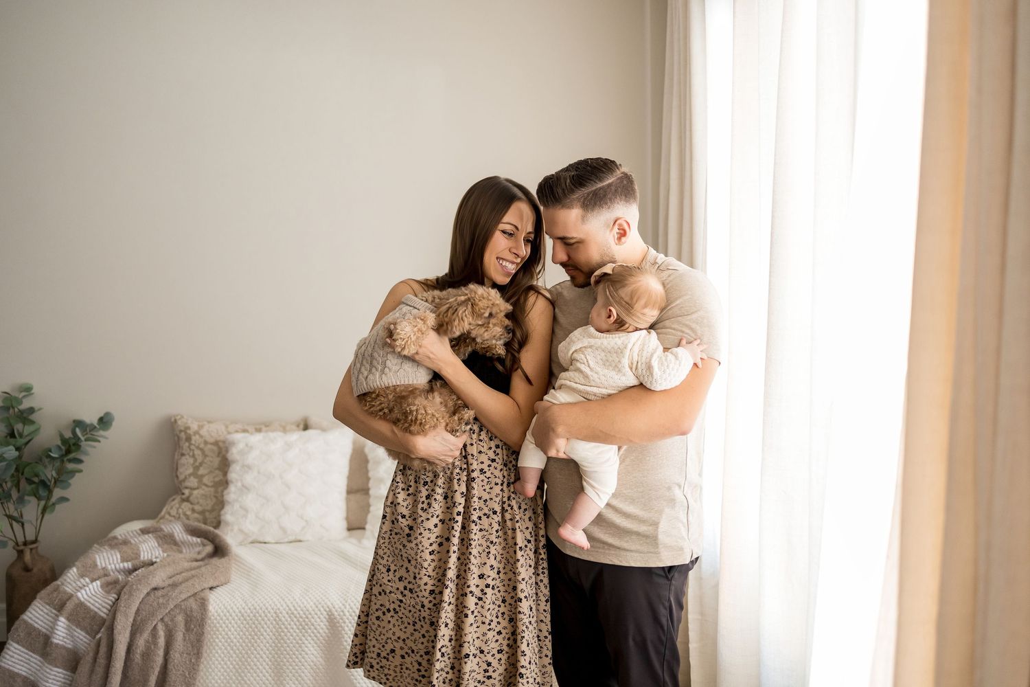 Young couple standing by window holding their newborn baby and small dog in a bright, cozy bedroom.