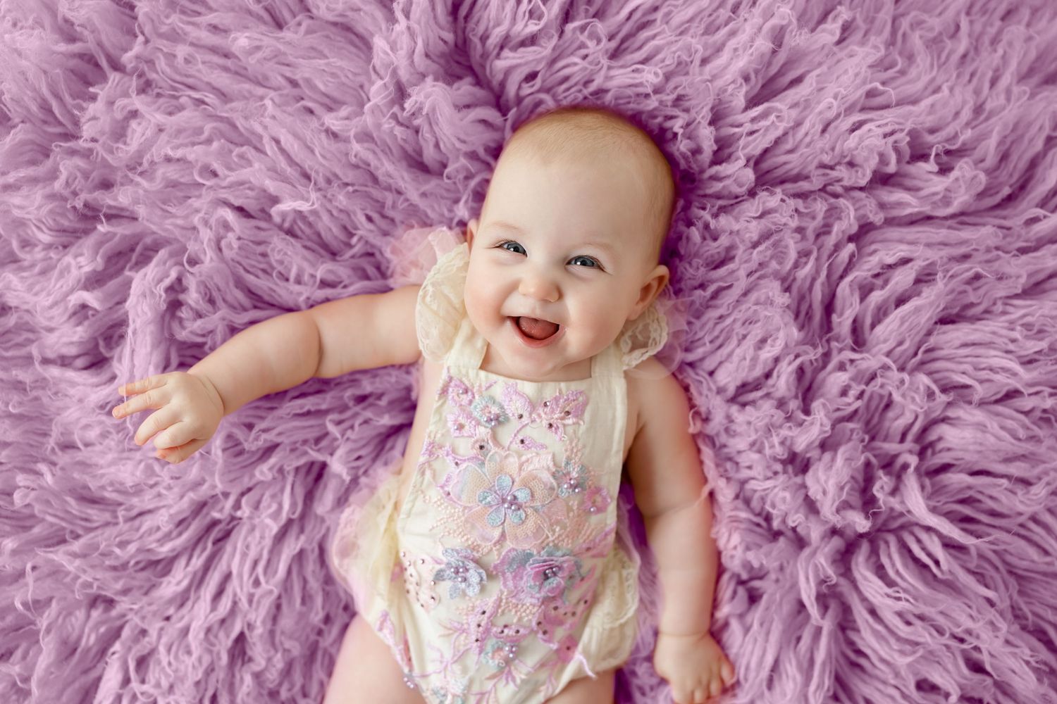 Baby in floral outfit lies on plush purple carpet during photo session.