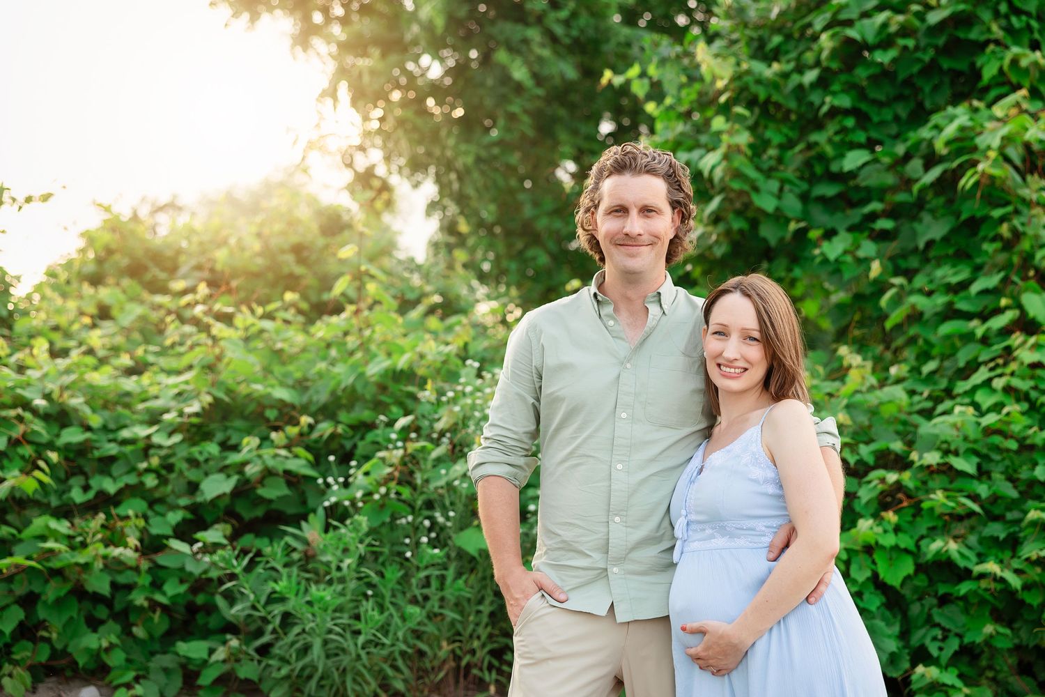Couple embraces during sunset maternity photo session surrounded by green foliage.