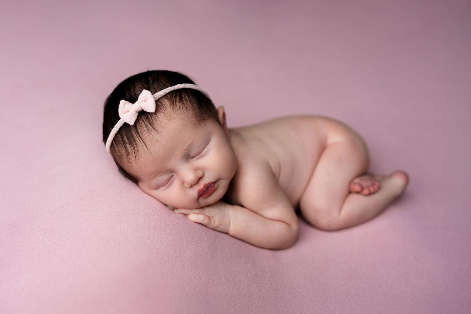 A sleeping newborn wearing a white bow headband rests peacefully on a pink background.