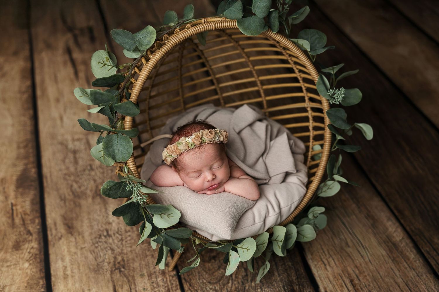 Sleeping newborn in basket surrounded by green eucalyptus leaves on wooden floor.