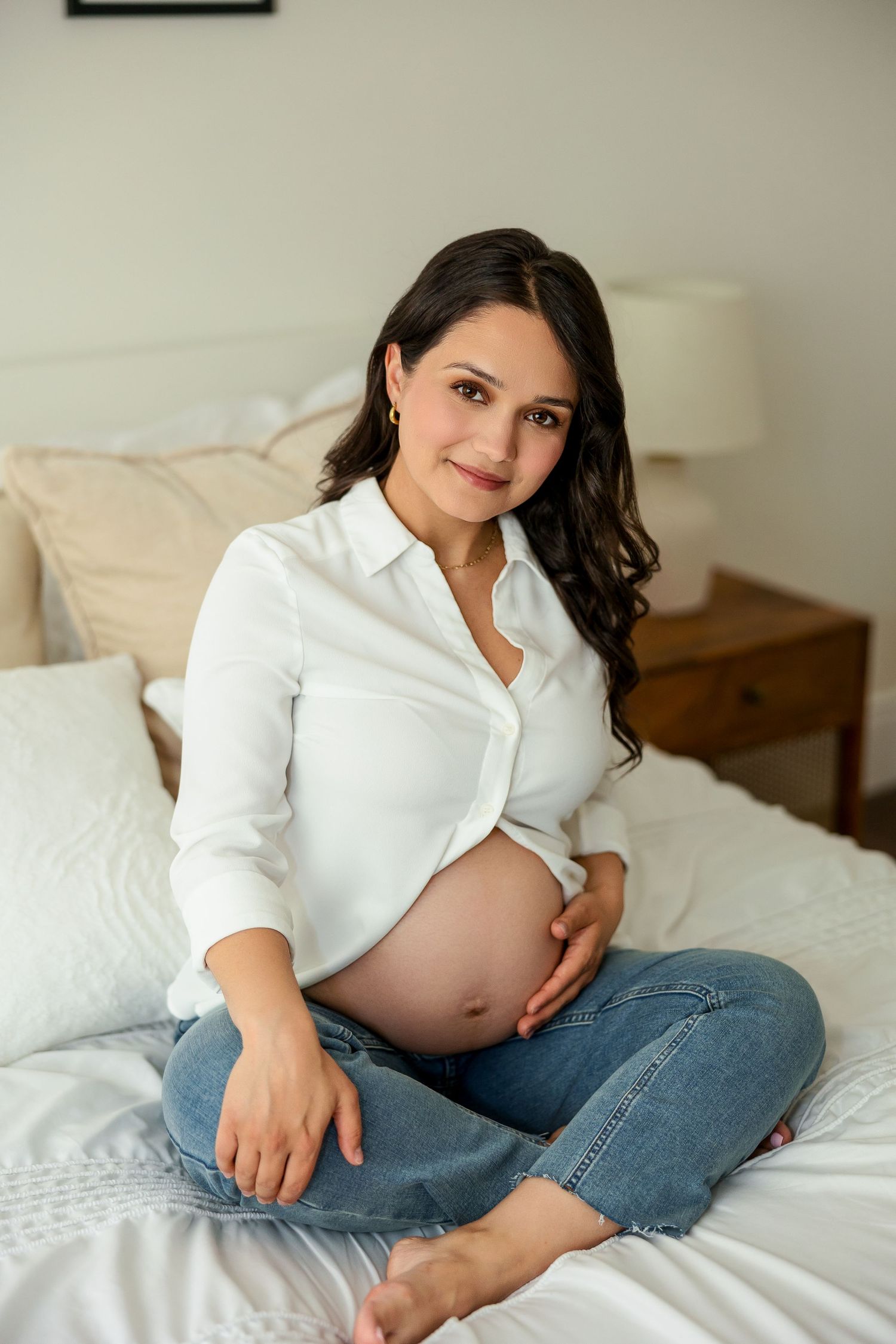 A pregnant woman in a white button-down shirt and jeans sits comfortably on a bed with white linens and pillows.
