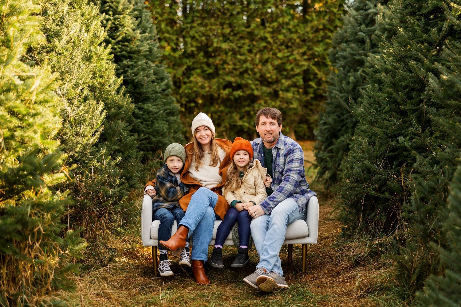 A family sits together on a white couch surrounded by evergreen trees at a Christmas tree farm.