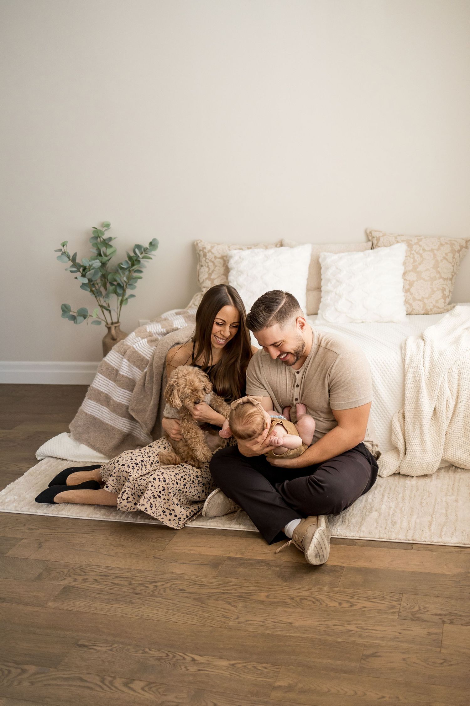 A young family with a newborn baby and golden doodle dog sitting on a rug against a bed in a cozy room.
