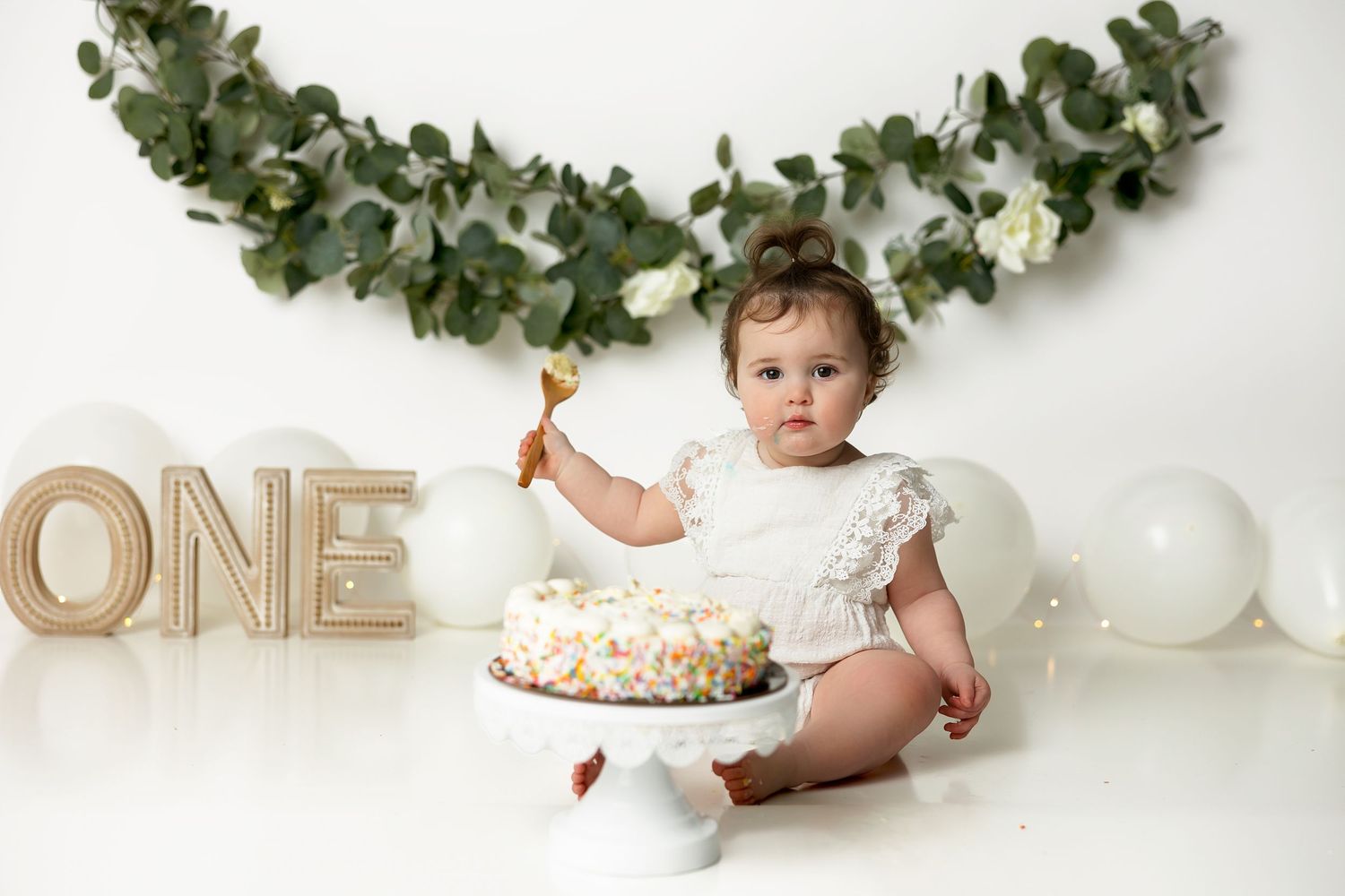 First birthday cake smash photo with white balloons, floral garland and light wood ONE letters against white backdrop.