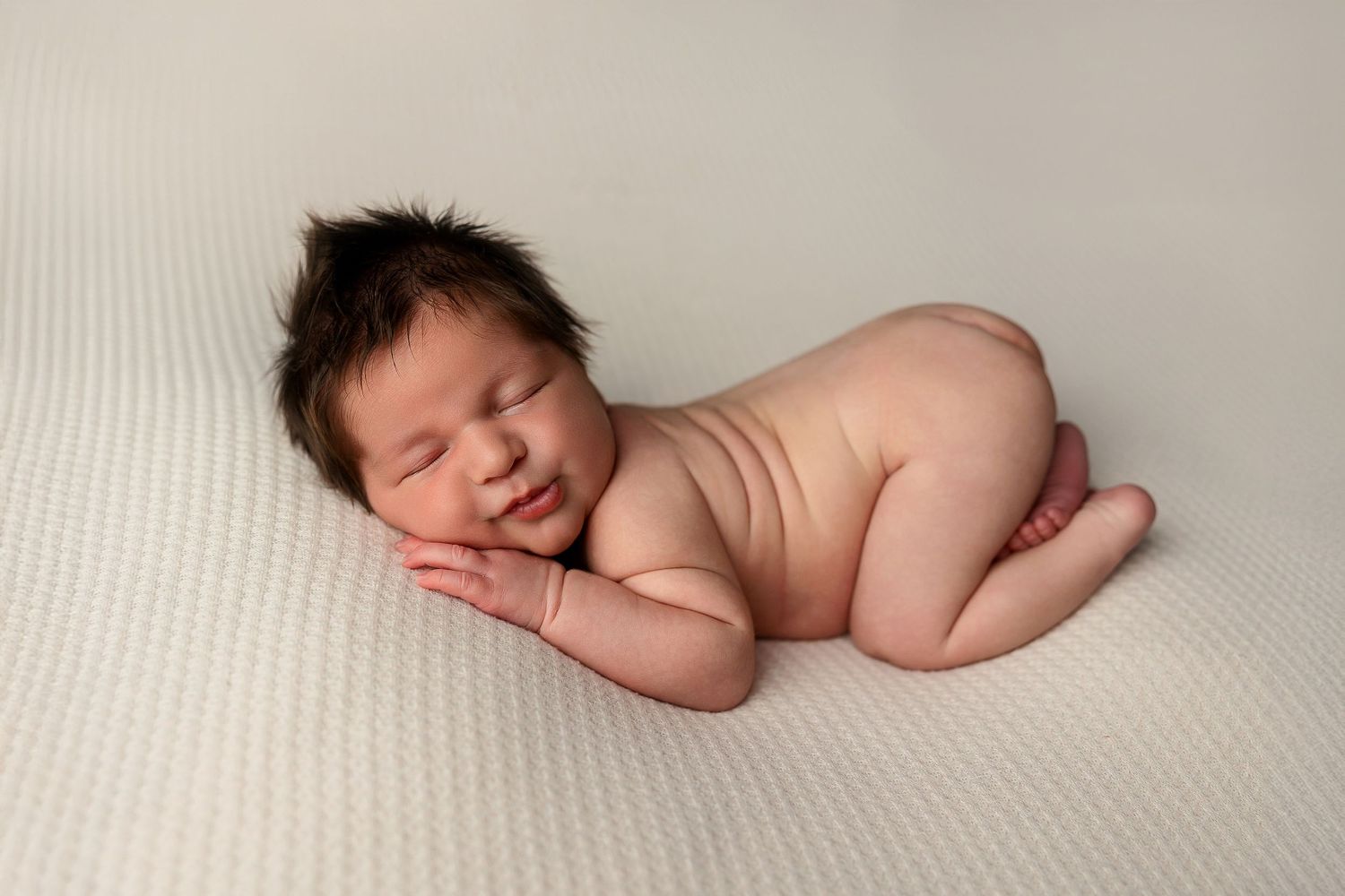 A sleeping newborn baby lies peacefully curled up on a soft white textured blanket in natural light.