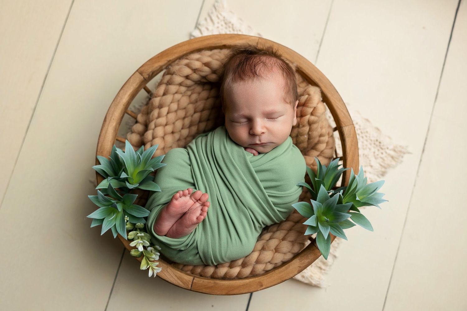 Newborn baby wrapped in green fabric with succulent decorations in wooden basket.