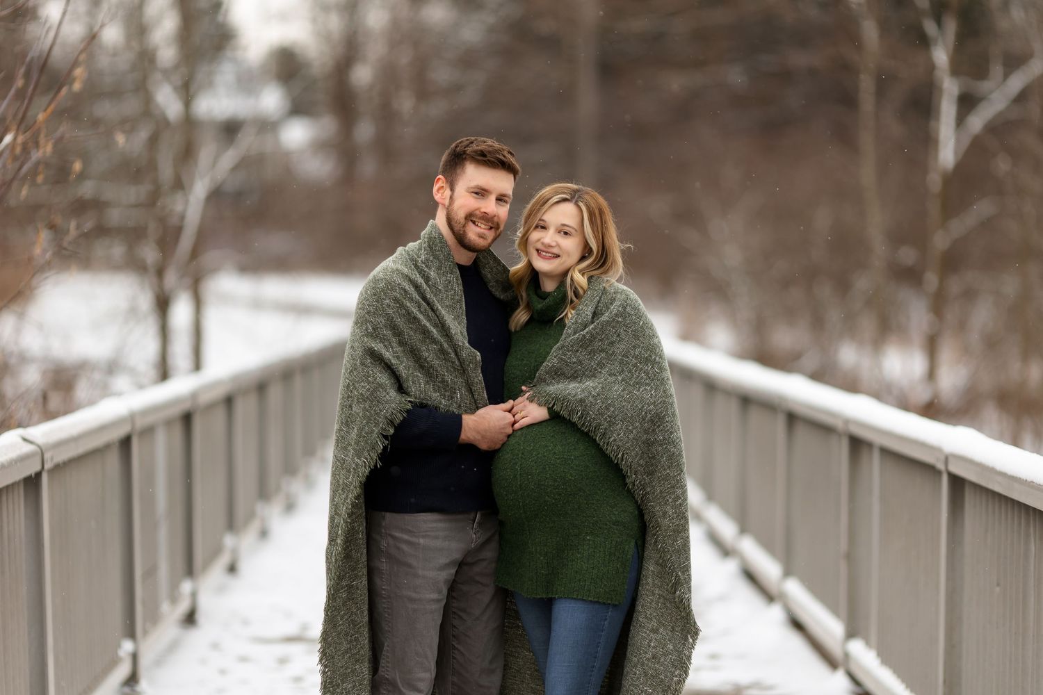 Couple wrapped in blanket standing on snowy bridge during winter photo session.