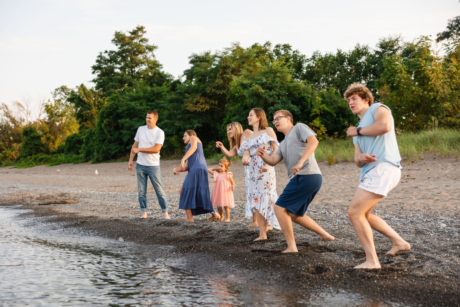Group of people in casual clothes playing and walking along the shoreline at sunset.