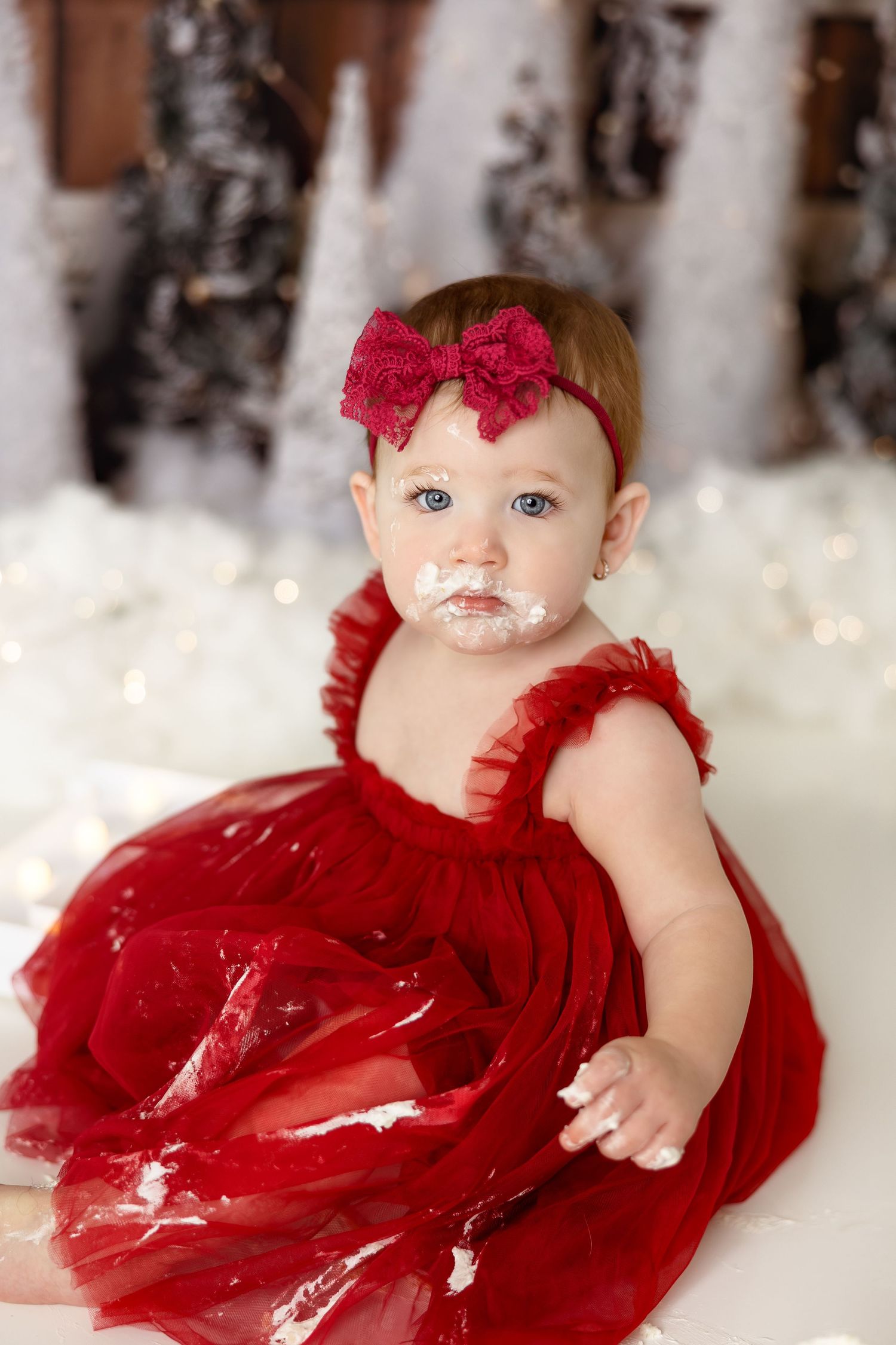 Baby wearing red tulle dress with bow sitting on white snowy floor with sparkly bokeh lights in background.