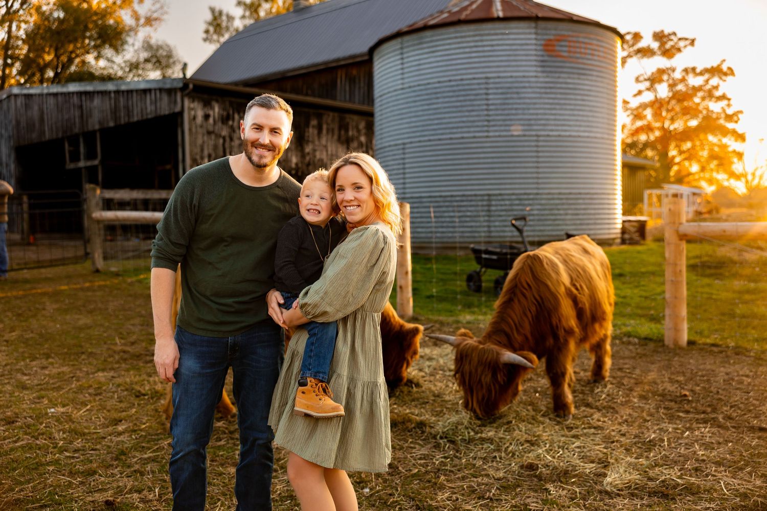 A couple poses by a metal grain silo and barn at sunset with a brown cow grazing nearby.
