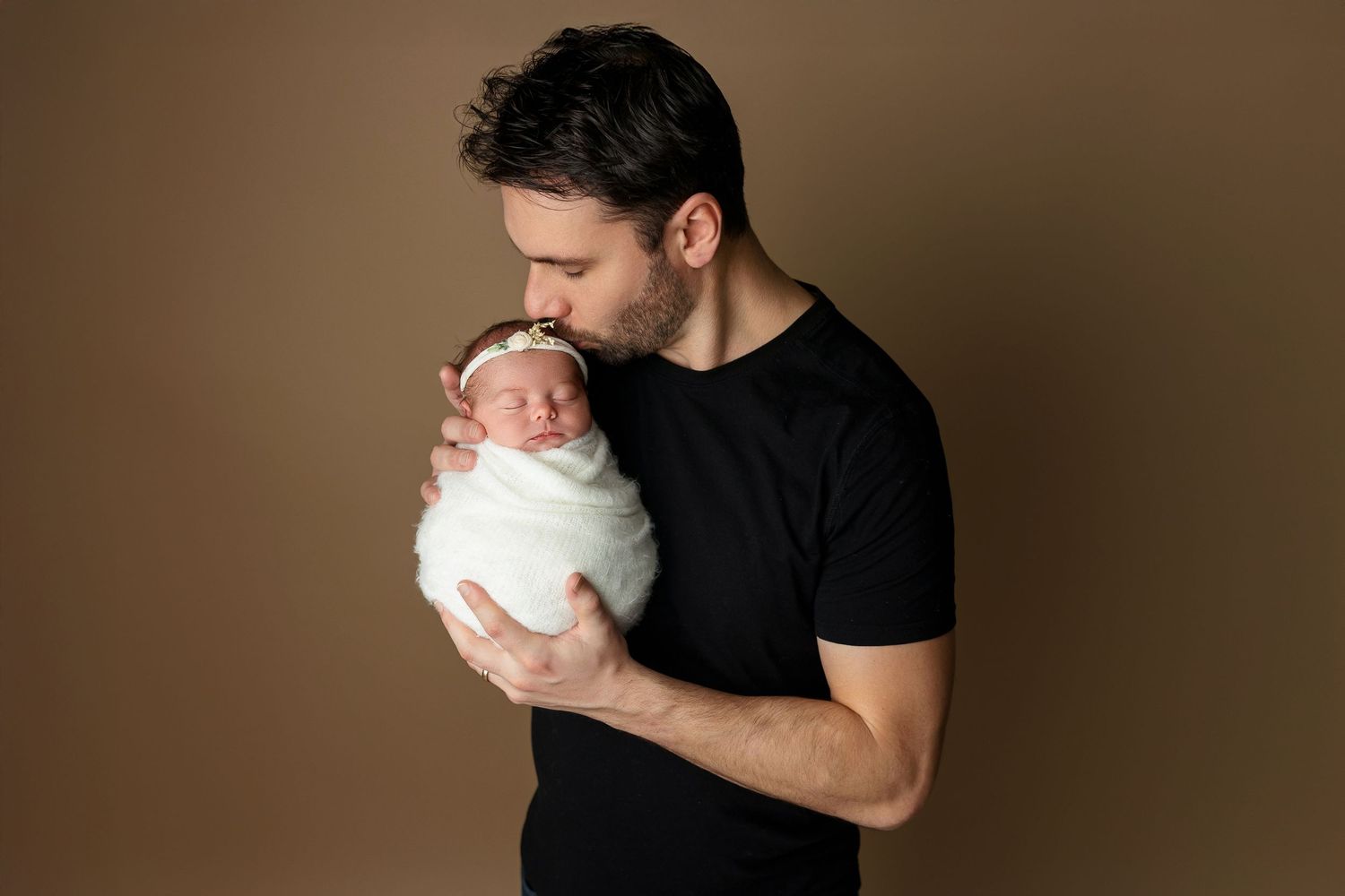 Tender moment between parent and swaddled newborn against brown backdrop.