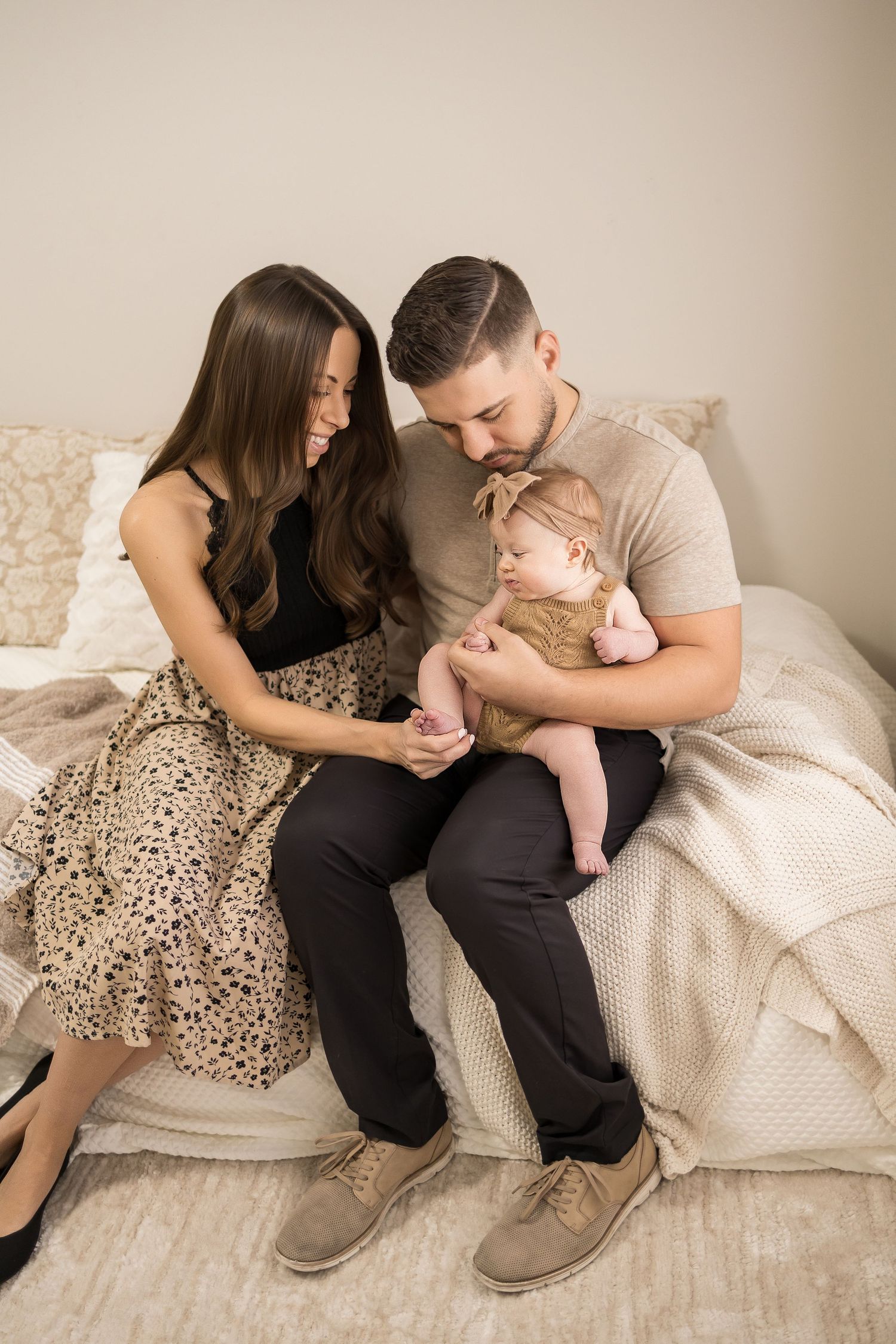 A young family sits on a bed, parents lovingly gazing at their newborn baby dressed in a tan outfit.