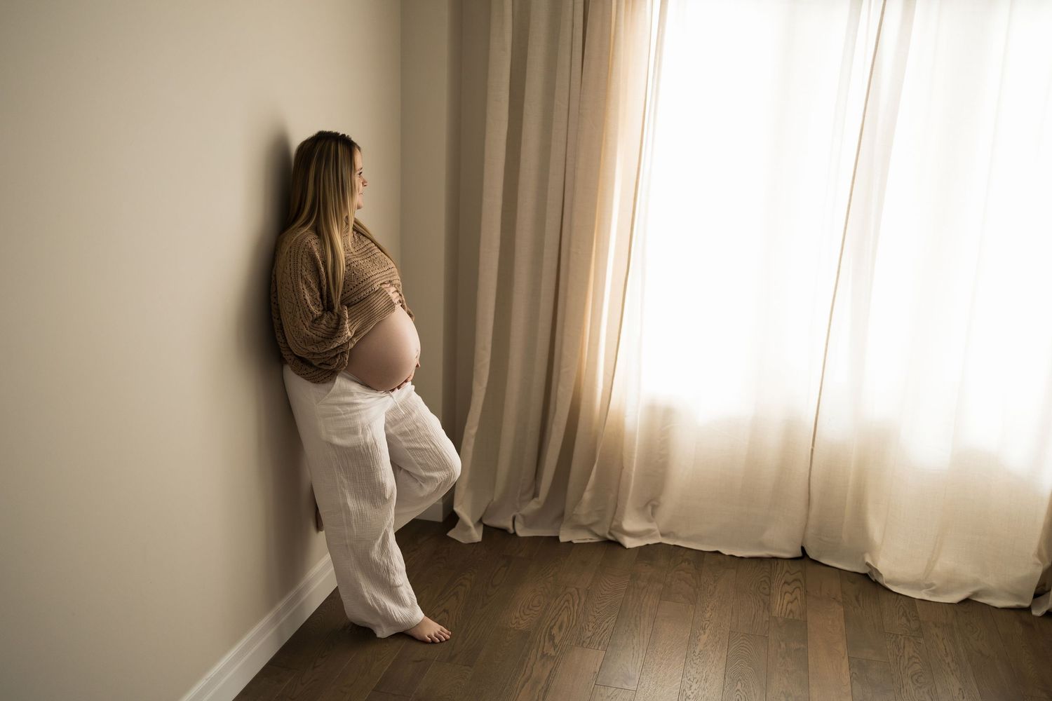 A person sits on a windowsill in a bright room with white curtains creating a contemplative silhouette.