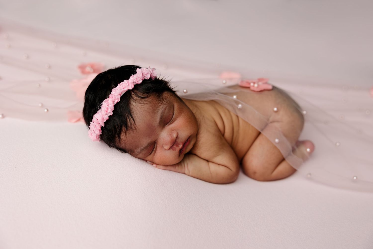 A sleeping newborn baby wearing a pink headband lies on soft pink fabric in a peaceful pose.