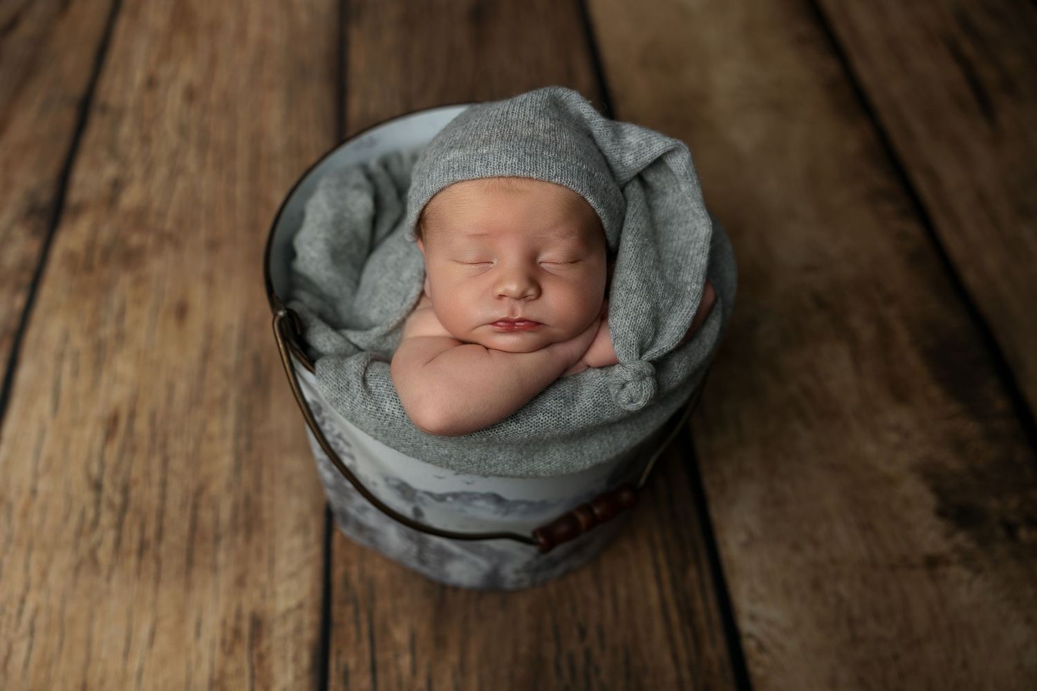 Peaceful sleeping newborn baby wrapped in soft gray blanket posed in wooden bowl on rustic wooden floor.