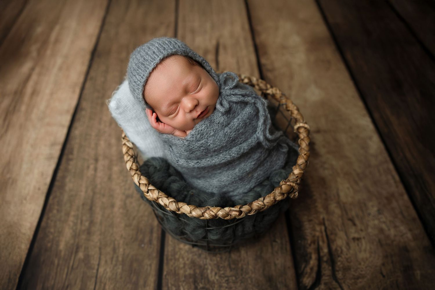 Newborn baby swaddled in gray knit wrap and bonnet sleeping peacefully in a woven basket on rustic wood floor.