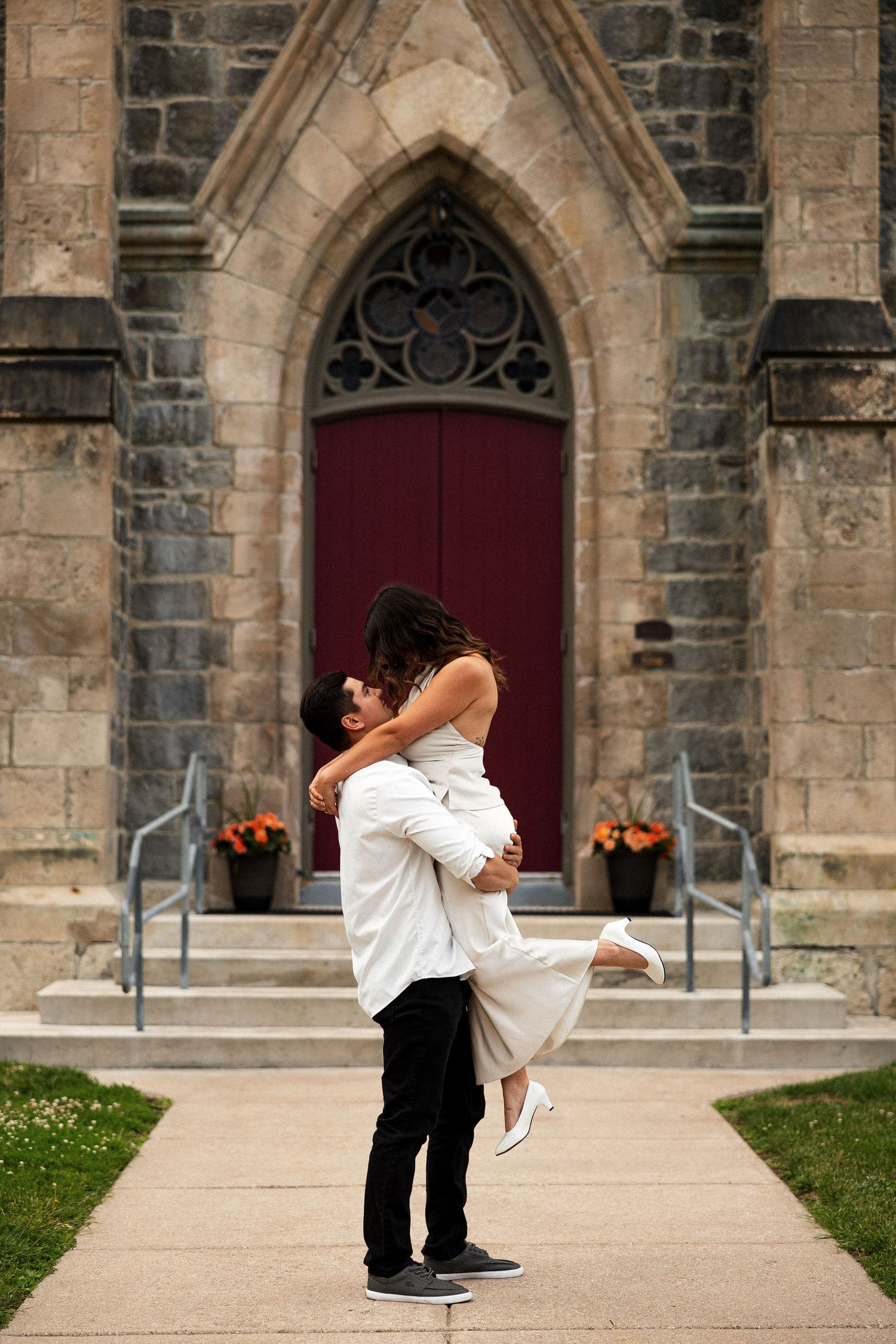 Couple embracing in front of a stone church with a red door, man lifting woman in a romantic pose.