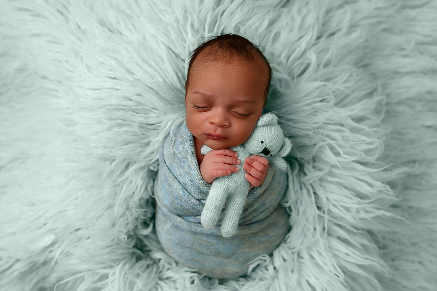 Sleeping baby in gray wrap cuddling small stuffed animal on fluffy white fur.