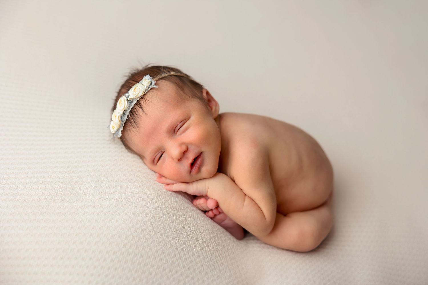 Sleeping newborn in curled pose with lace headband on white textured backdrop.