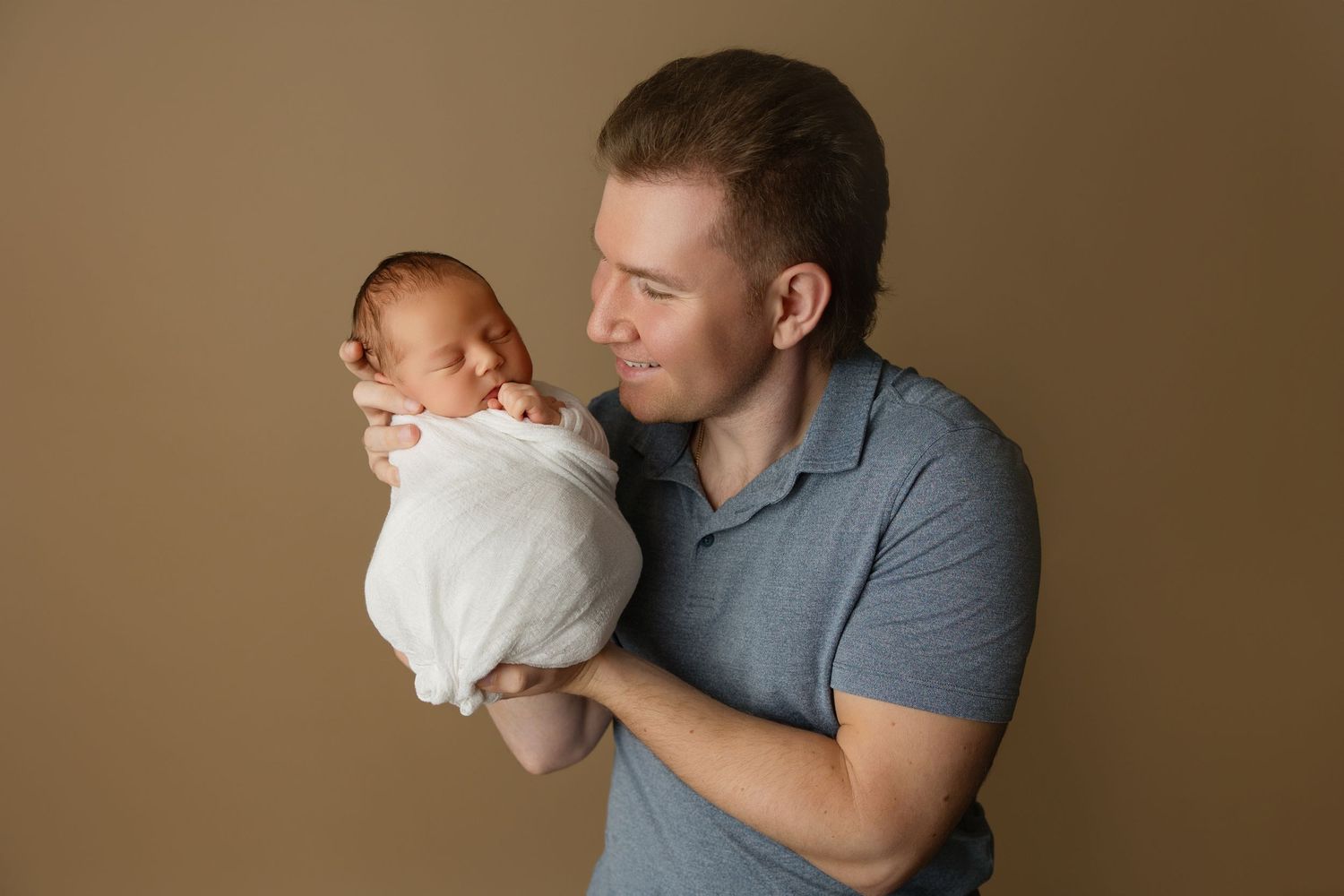 Studio portrait of parent in grey polo shirt holding newborn wrapped in white.