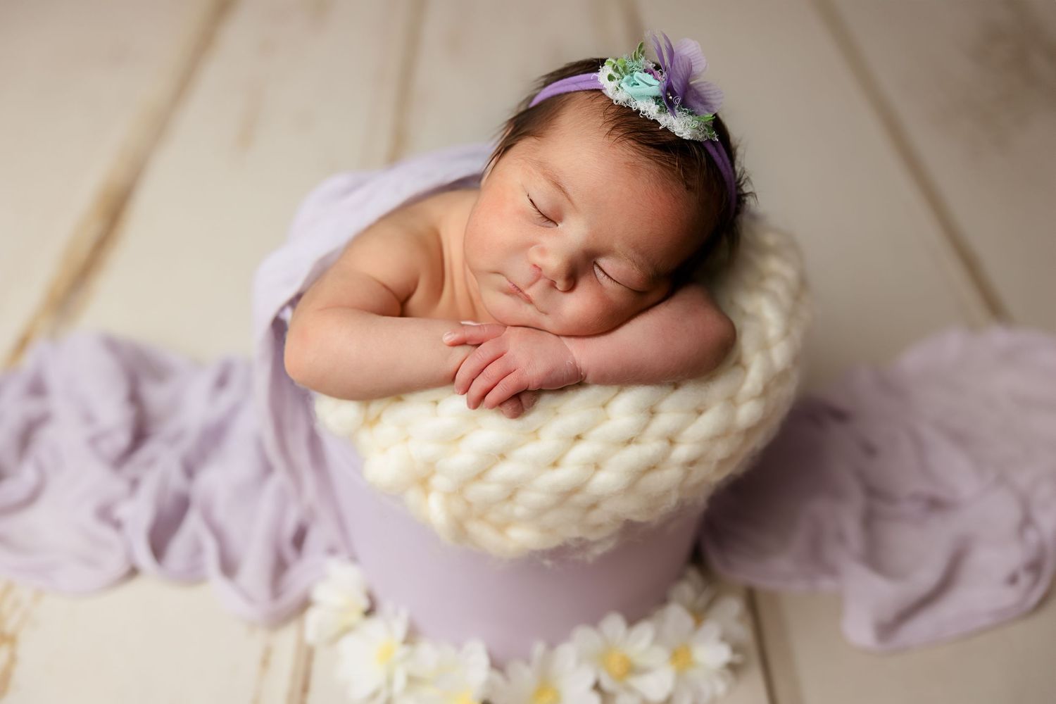 Sleeping newborn posed in knitted cream bowl with purple accents and flower headband.
