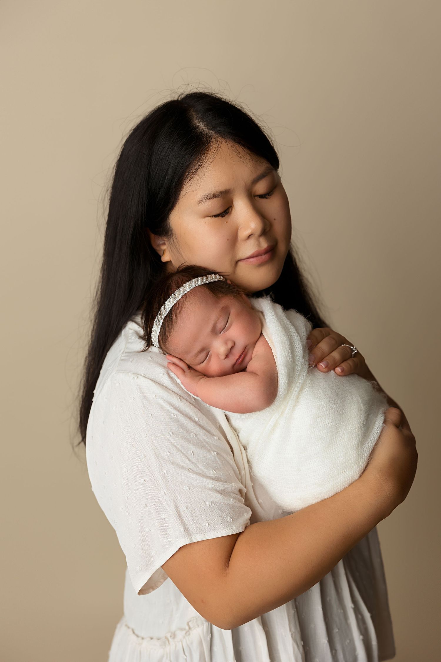 Mother in white dress holding sleeping newborn wrapped in white against beige background.
