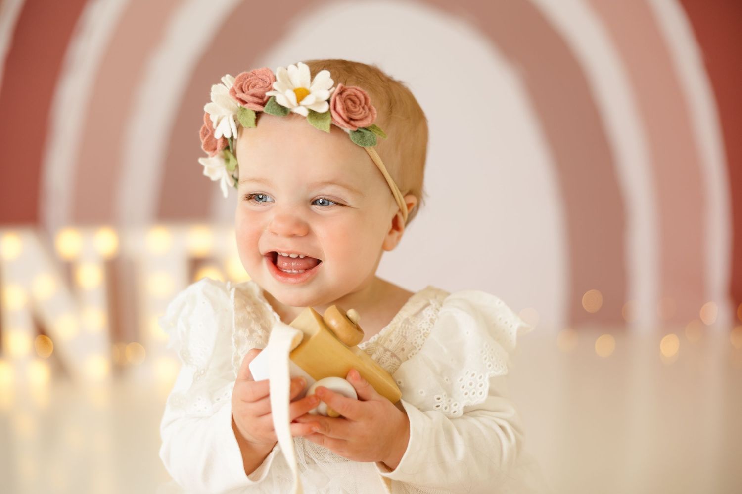 Smiling baby in white dress and floral headband against pink circular backdrop with 'ONE' letters.