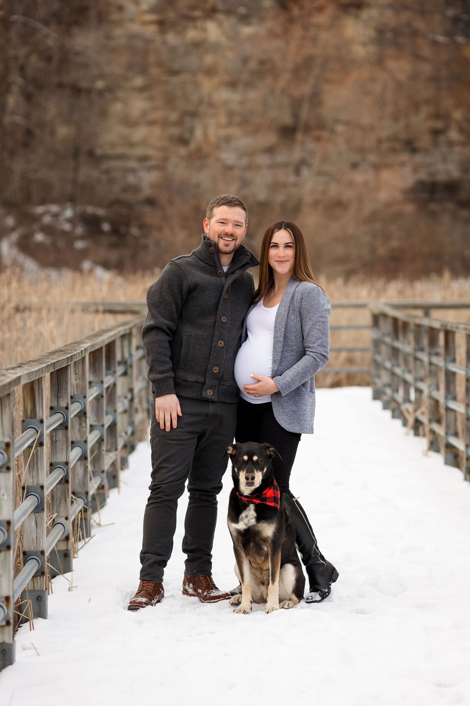 Expectant couple poses with their dog on snow-covered bridge during maternity session.