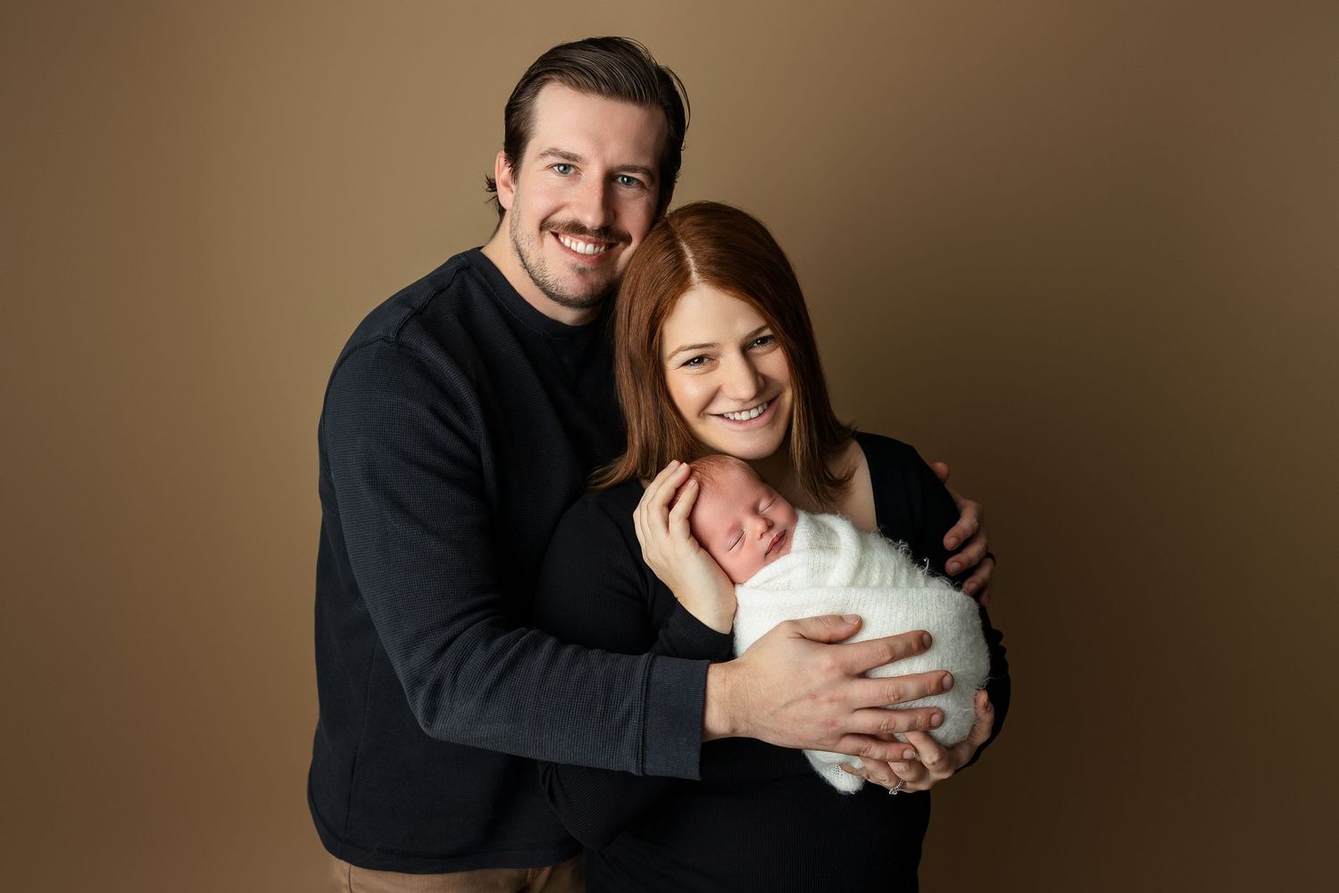 Couple in dark sweaters embraces while holding newborn wrapped in white.