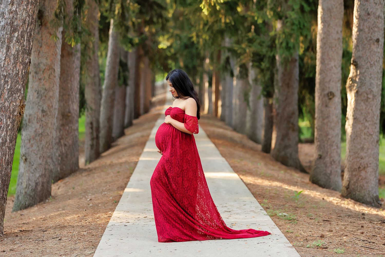 Woman in flowing red dress poses along tree-lined path during outdoor maternity photoshoot.