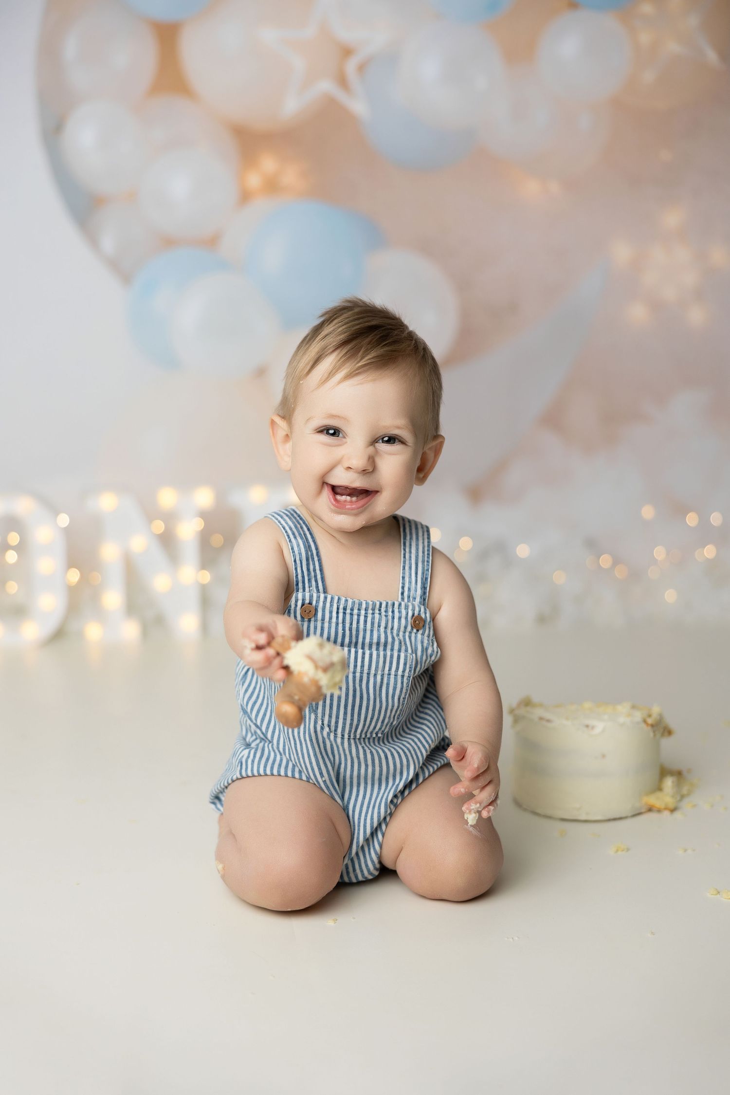 Baby wearing blue striped romper sitting near birthday cake setup with twinkle lights and balloons.