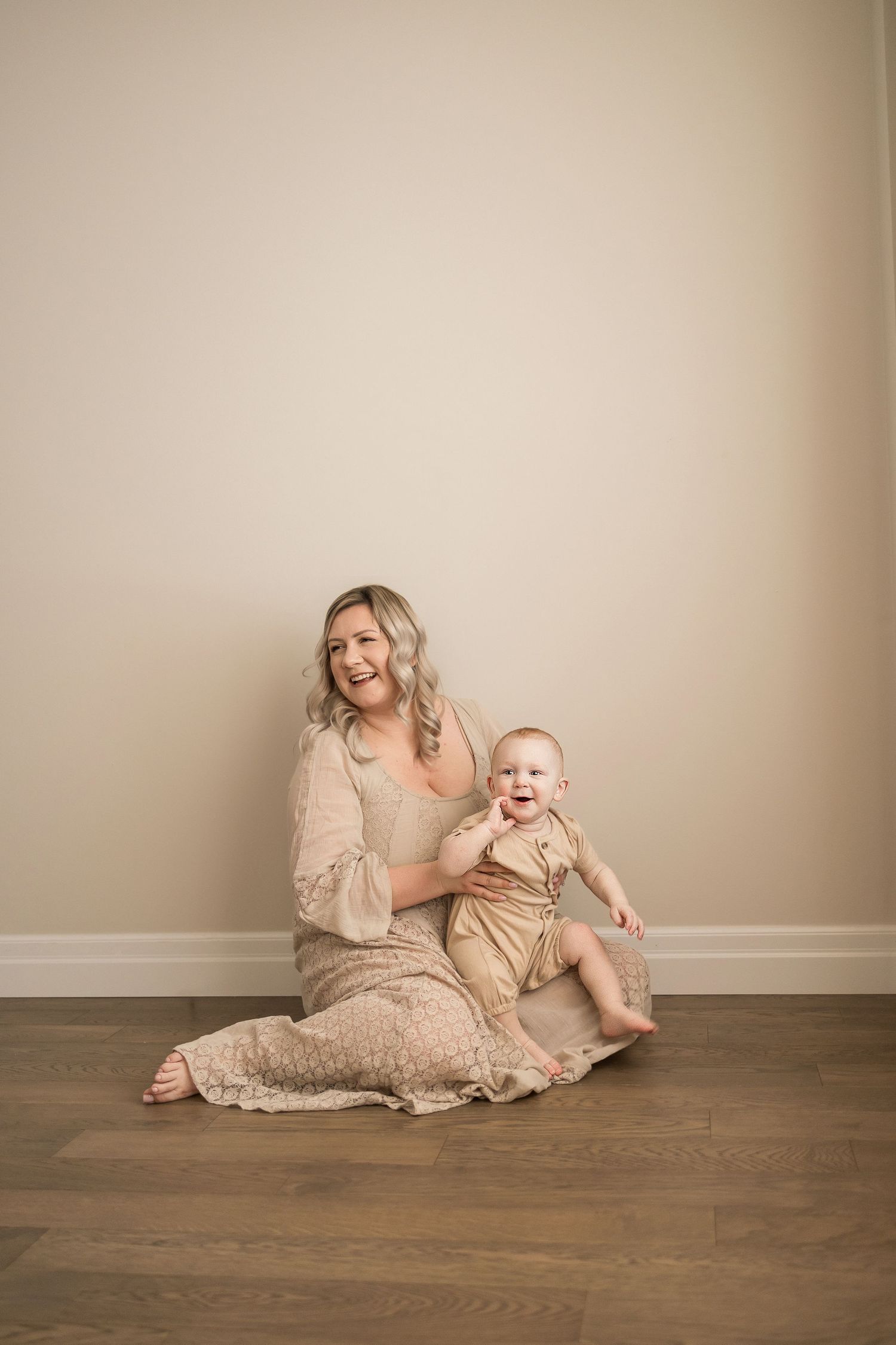A tender portrait in neutral tones shows a parent and baby sitting together on hardwood flooring against a beige wall.