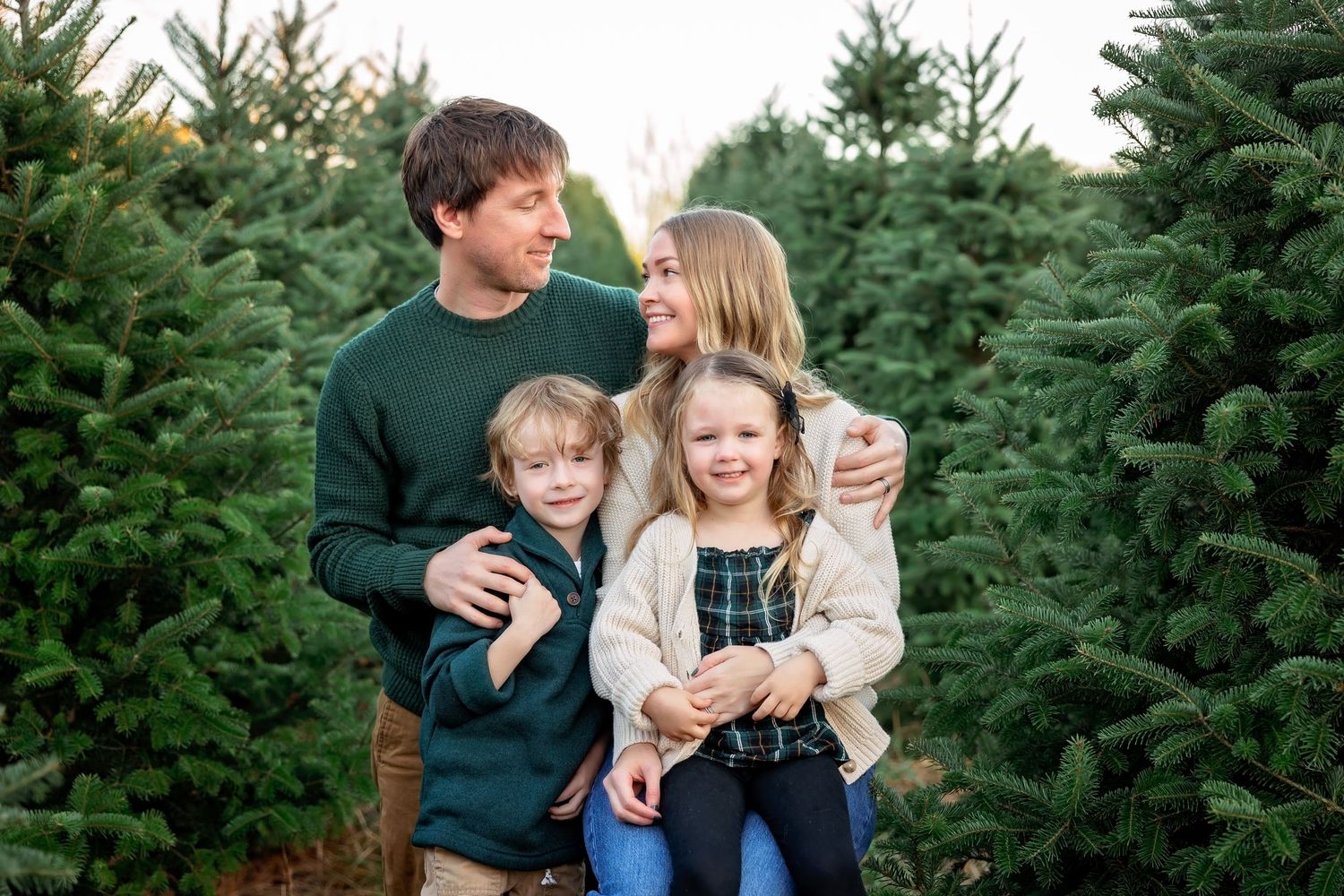 A loving family poses together among evergreen Christmas trees at a tree farm.
