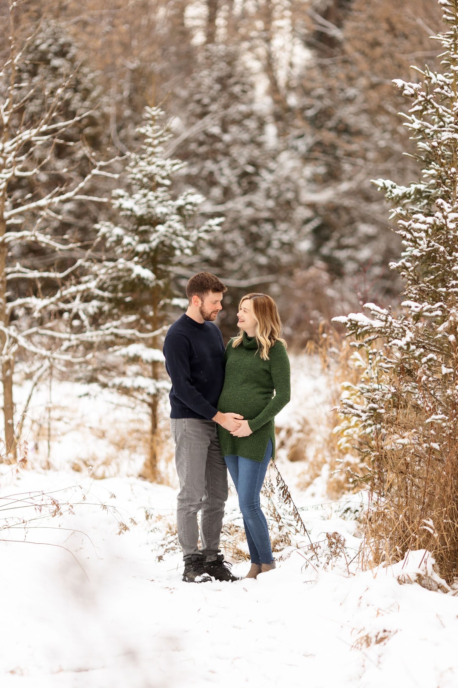 A romantic winter maternity photo with a couple standing among snow-covered branches.