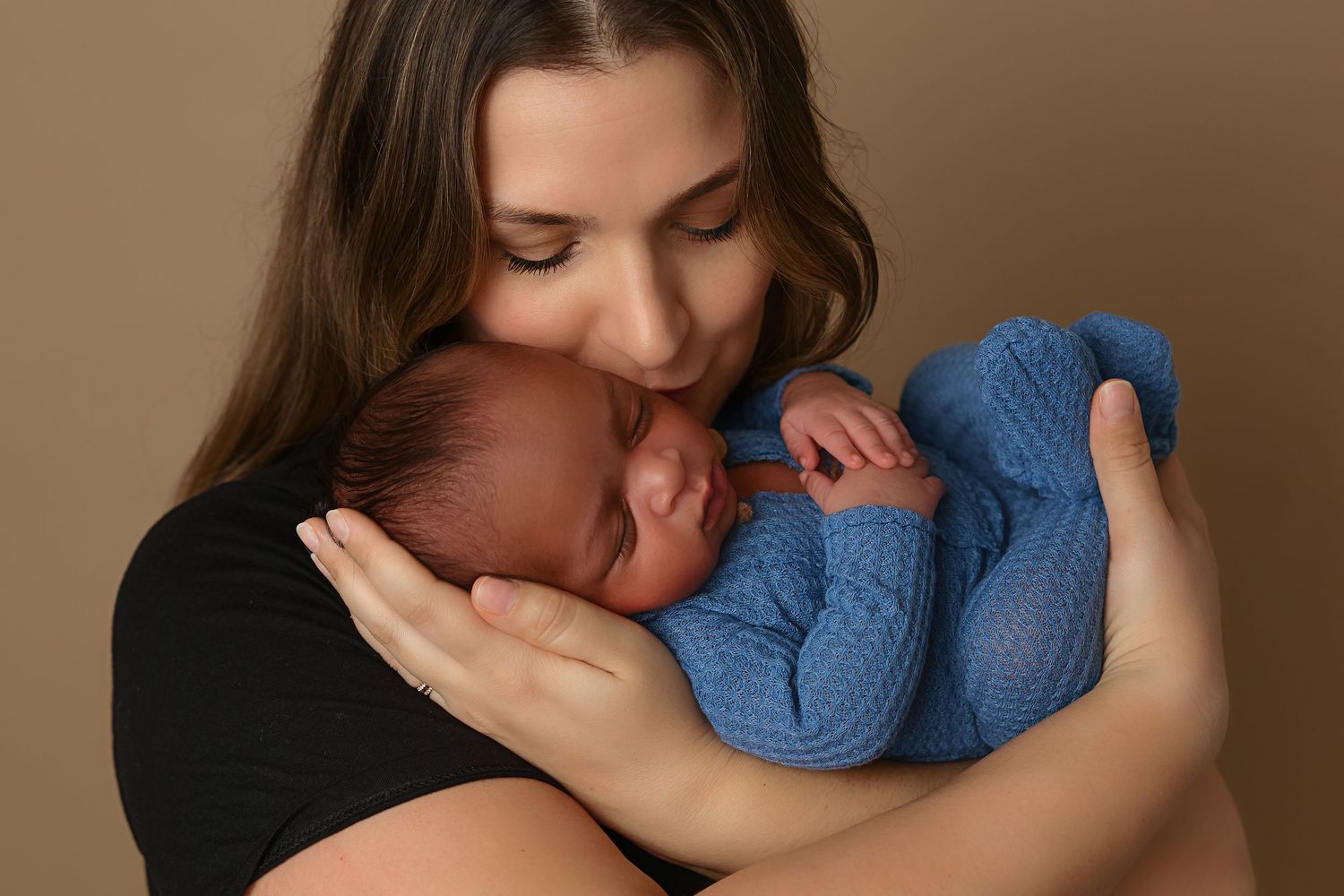 Mother gently cradling newborn wearing blue against warm brown studio background.