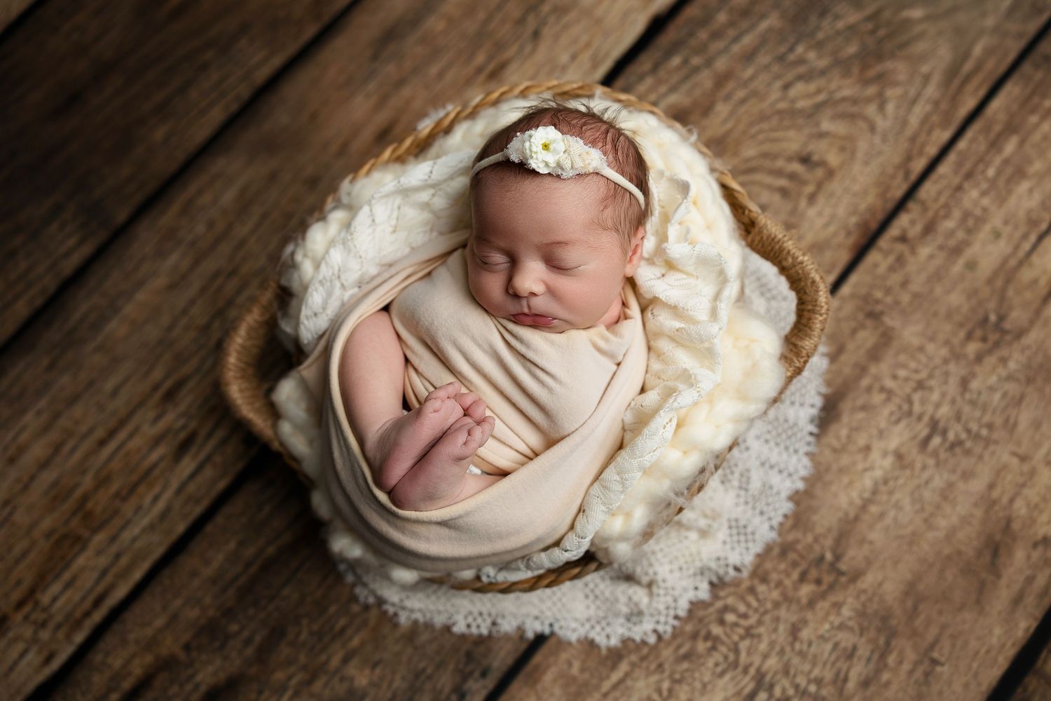 Newborn baby sleeps peacefully in cozy white wrap inside wooden bowl on rustic floor.