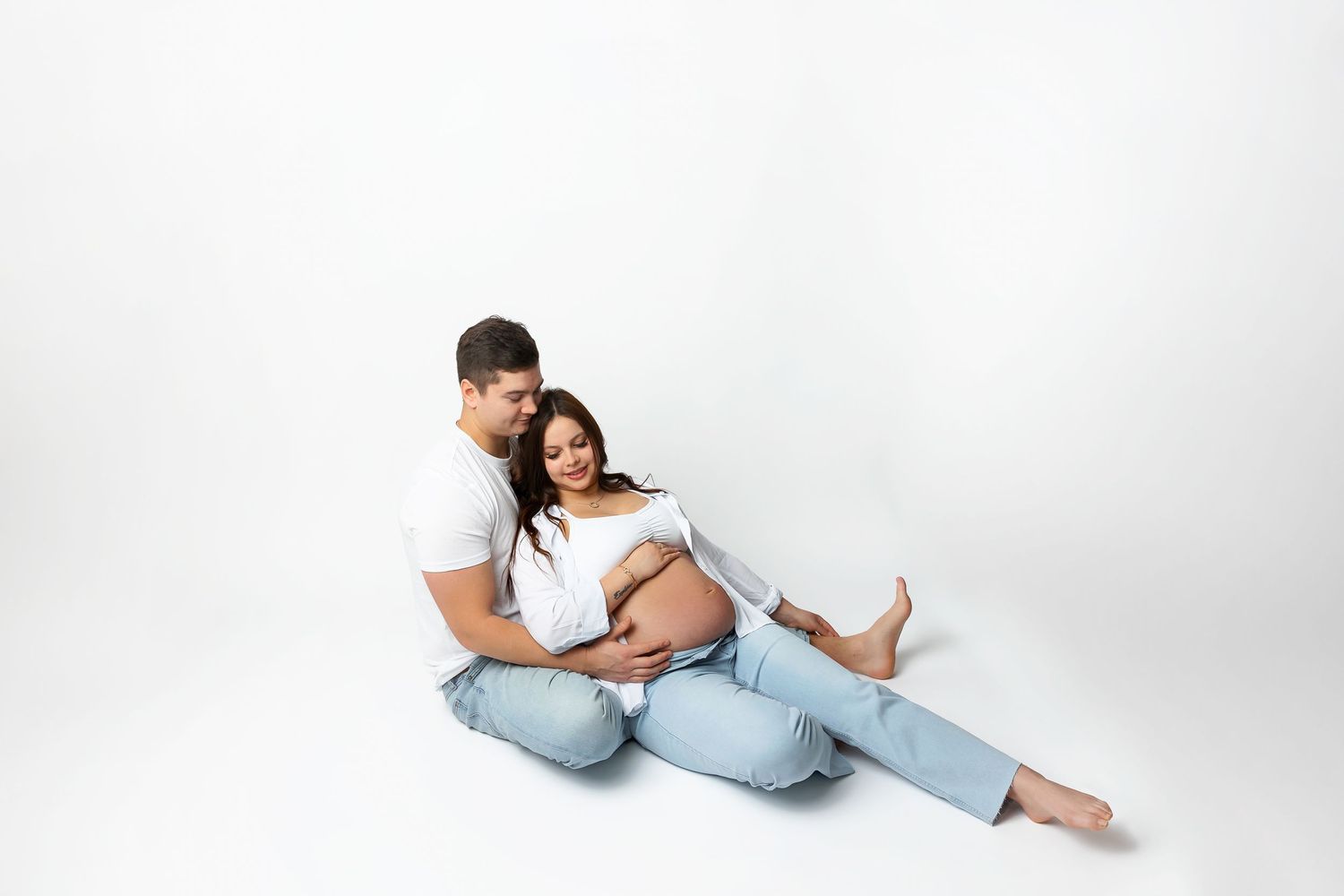 Couple in casual attire sitting on white backdrop during maternity photoshoot.