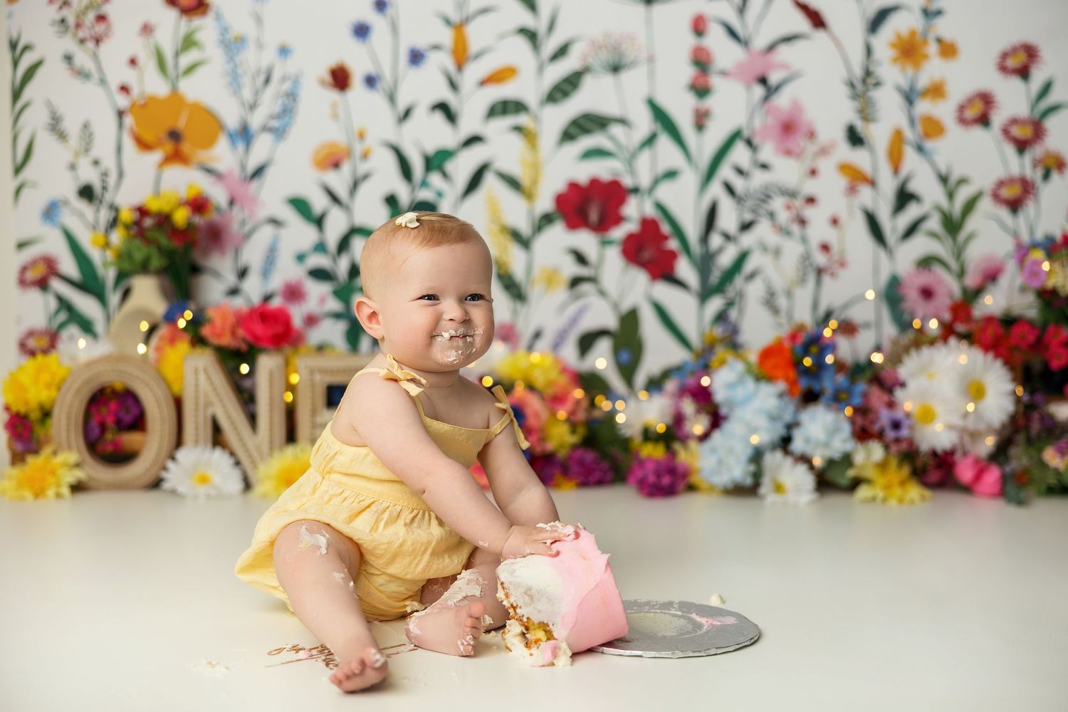 Baby in yellow romper lies on floor surrounded by colorful floral backdrop during photo session.
