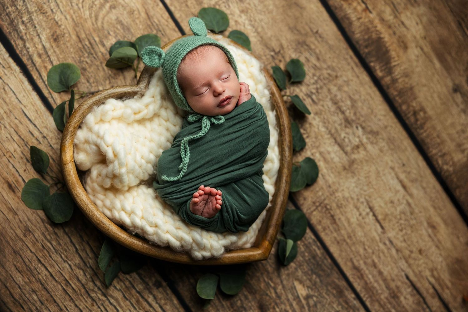 A sleeping baby in green swaddle rests on rustic wooden surface with eucalyptus leaves.