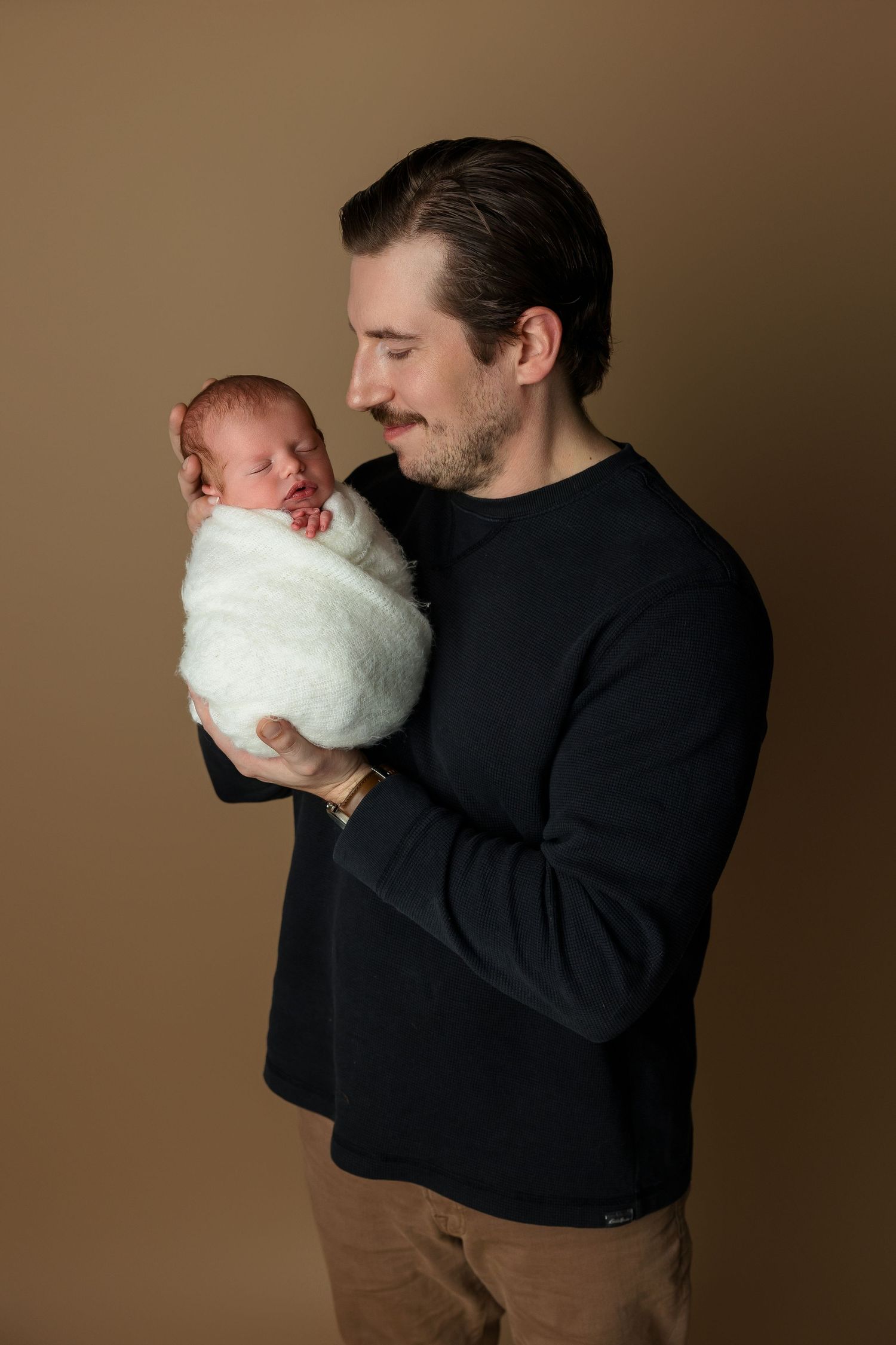 Intimate moment between parent and swaddled newborn against brown backdrop.